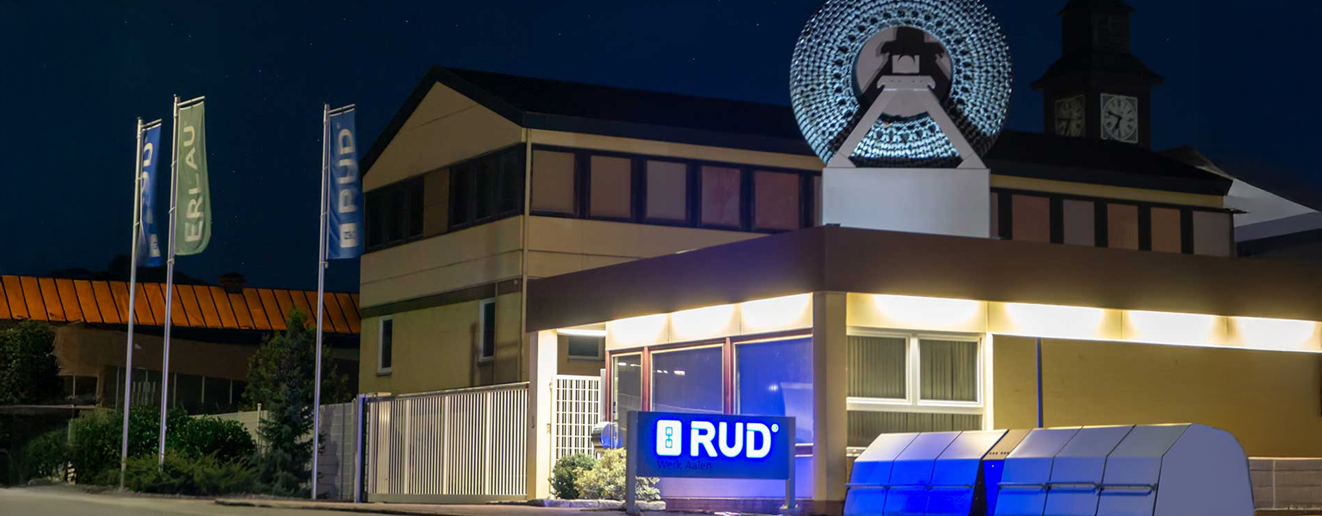 Das Gebäude von RUD bei Nacht, beleuchtet mit dem Firmenlogo und Fahnen der Marken "ERLAU" und "RUD" vor dem Werk in Aalen.